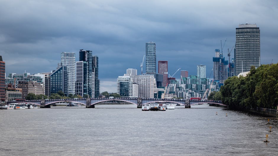 A panoramic view of a city skyline across a river, featuring numerous modern high-rise buildings with glass facades, some under construction with cranes visible, and other mid-rise structures. The river in the foreground has several white boats and barges docked near the banks, with a stone bridge spanning across the water. On the right side, dense green trees line the riverbank, while on the left, urban buildings are situated close to the water’s edge. The sky above is filled with dark, heavy clouds indicating overcast weather. This image captures a vibrant urban scene that could relate to a home relocation or moving logistics in a city environment, suitable for a page about removals in Lambeth, with a focus on transport and packing considerations associated with furniture transport and moving processes.