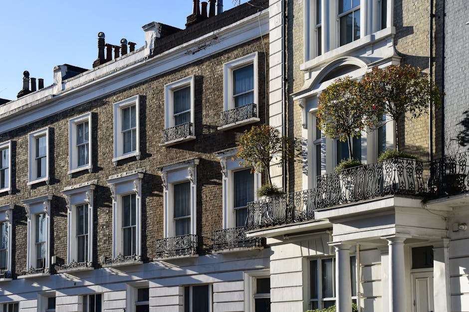 A row of terraced houses with brick and stone facades, large front windows, and pitched roofs is visible beyond a large, well-maintained green lawn. Several tall, leafless trees are situated along the line of houses, with a few smaller trees that have some yellow-green foliage on the left side of the image. In the foreground, a few people are lying on or sitting near the grass, and others are walking or standing along the pavement adjacent to the houses. The sky above is clear with a few fluffy white clouds, indicating a bright and sunny day. The scene captures an outdoor residential area typical of urban house removals or home relocation settings, with natural lighting highlighting the details of the houses and the open space of the grass, suitable for a professional removals company like Man With a Van Lambeth handling furniture transport and packing in the Clapham Common area.