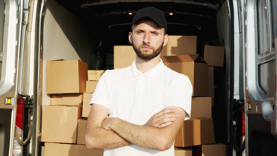 A man with a beard, wearing a red cap backwards, a checkered shirt, and a beige and brown jacket sits on the open back of a van used for home relocation. Behind him inside the van are several cardboar