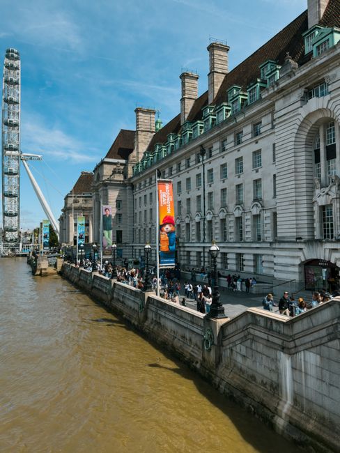 A view of a large historic white building with green roof details and multiple chimneys, situated along the River Thames in London. The building's facade features tall windows and decorative architect