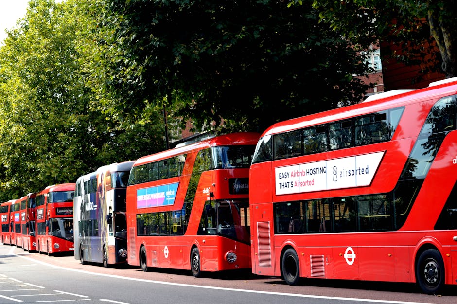 An aerial view of a city street with parked cars along the curb and moving vehicles on the road, including a red double-decker bus and several smaller cars, with pedestrians walking on the sidewalks. 